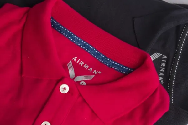 Fotografía desde arriba de una playera polo color rojo de la marca Airman doblada encima de otra playera color negro.